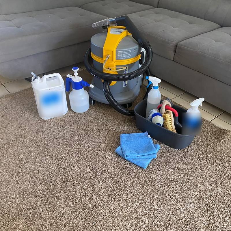 Machine de nettoyage professionnelle, pulvérisateurs et brosses prêts devant un canapé d’angle gris, sur un tapis