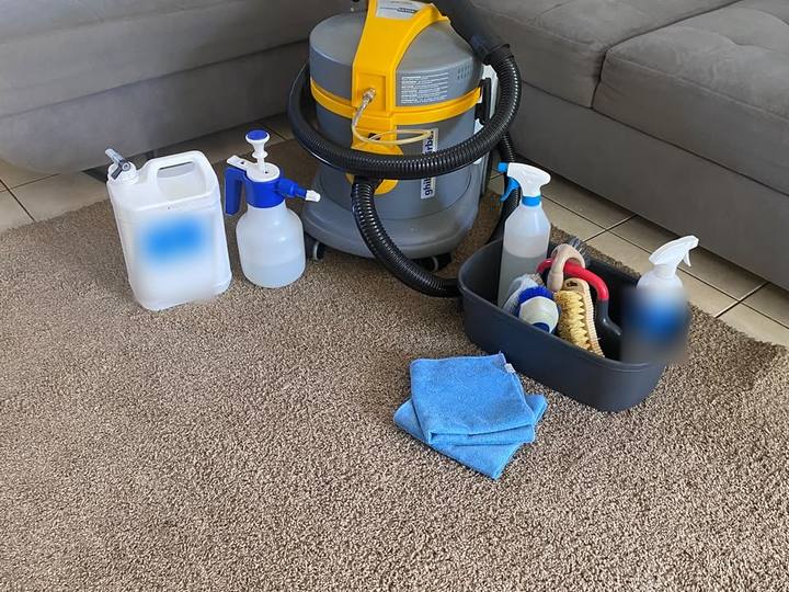 Machine de nettoyage professionnelle, pulvérisateurs et brosses prêts devant un canapé d’angle gris, sur un tapis