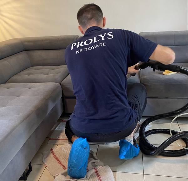 Intervenant Prolys passant la buse de la machine de nettoyage sur un canapé d’angle gris, sol protégé